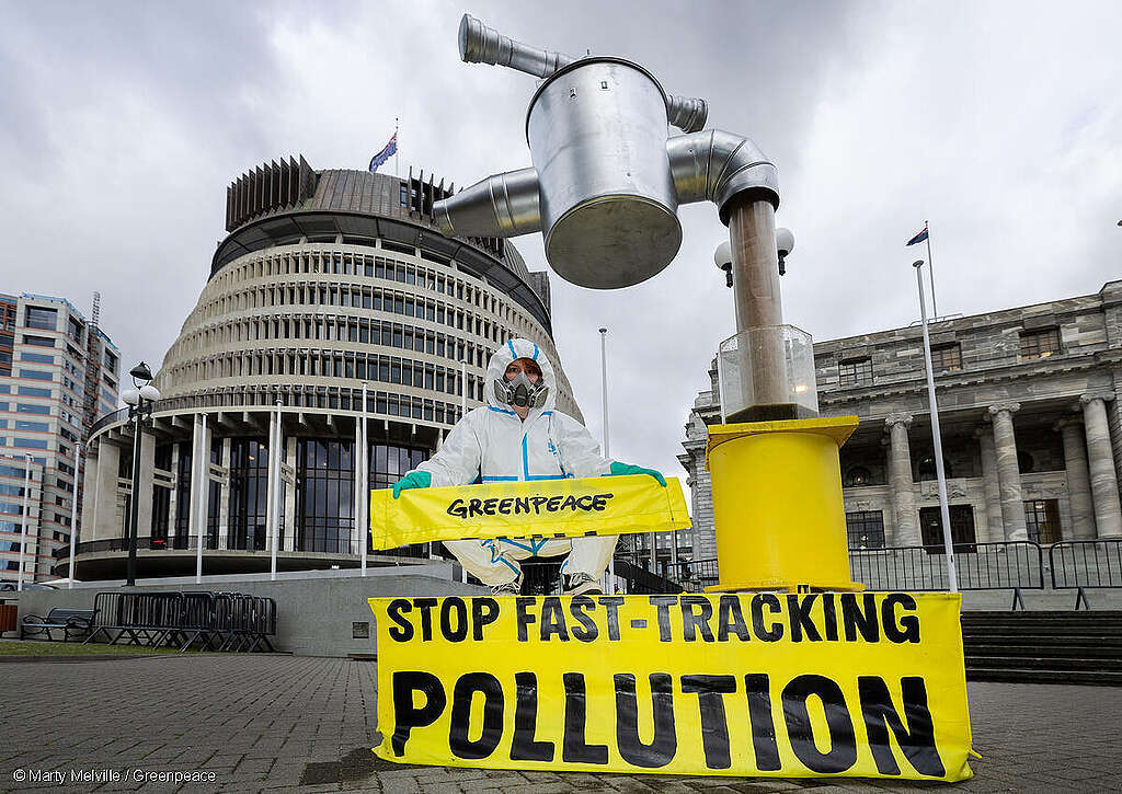 A giant tap with effluent pouring out, sits in front of the Beehive, Wellington. A Greenpeace activist, wearing a hazmatsuit holds a sign saying "Stop fast-tracking pollution"