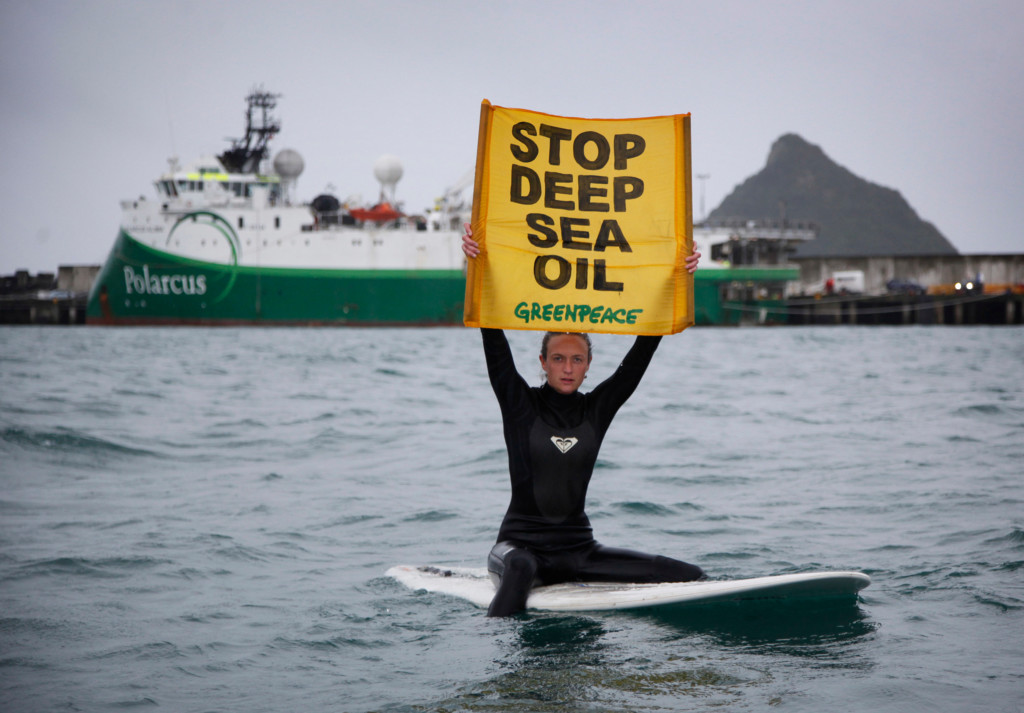 An activist on a surfboard in the ocean holds a yellow banner reading 'stop deep sea oil' in front of a large survey vessel at Port Taranaki