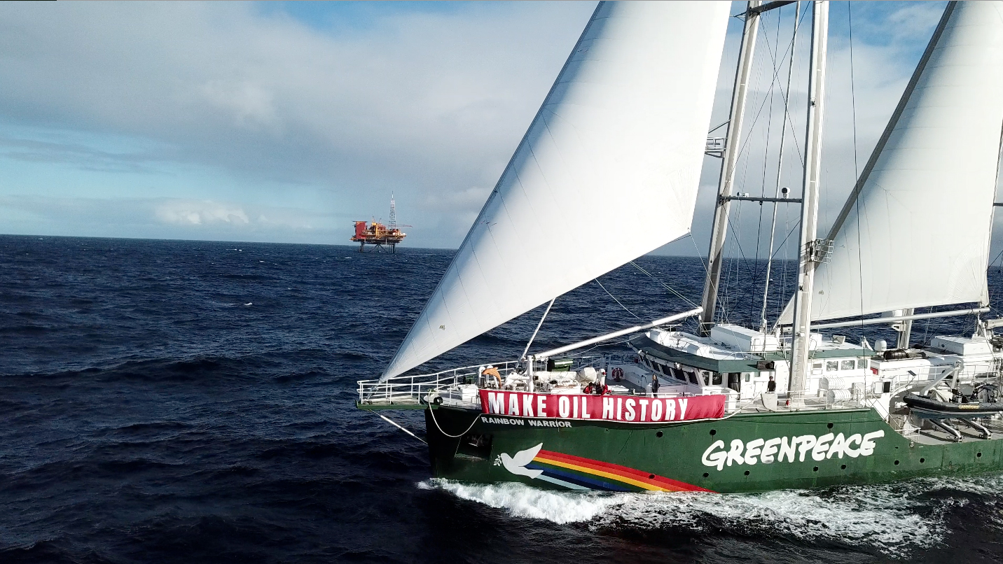 The Rainbow Warrior participates in a protest against oil drilling off the coast of NZ. Onboard is a banner reading 'make oil history'. In the background is an oil rig.