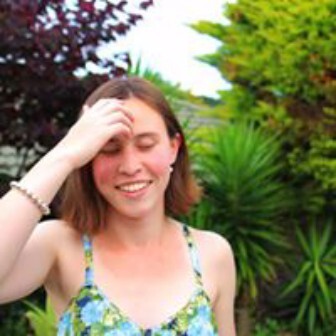 The author Áine Kelly-Costello stands smiling in the sun with green trees behind her.