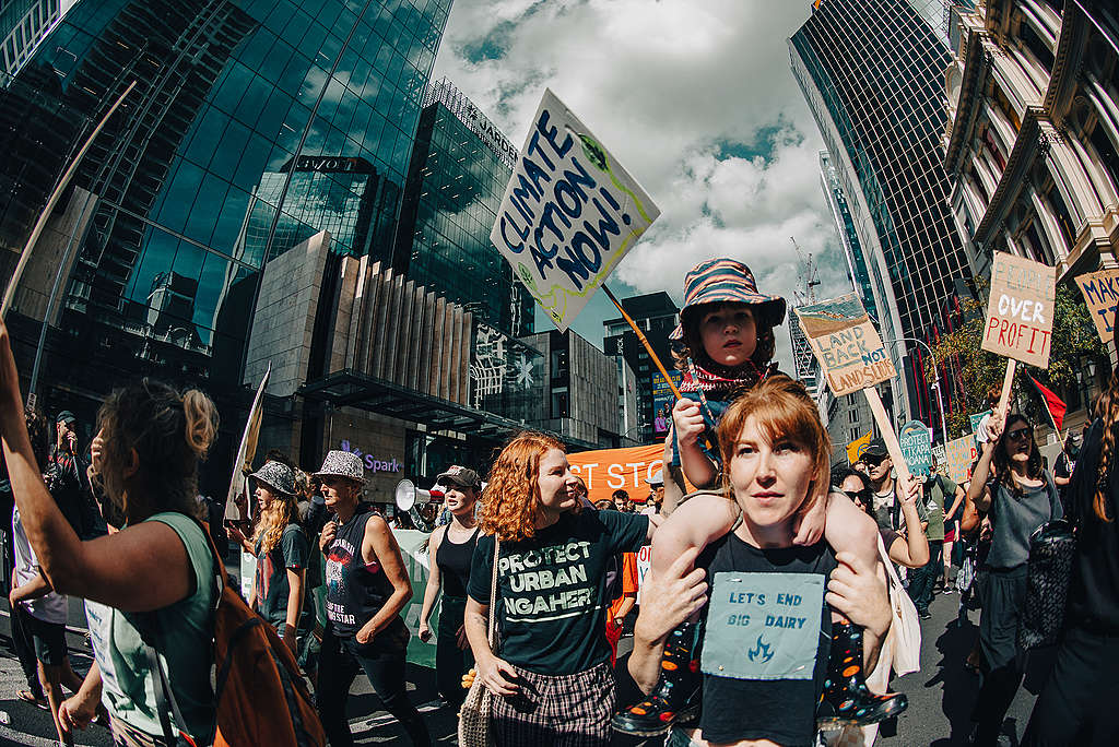 A child is sitting on a woman's shoulders, walking in street, they have a sign that says Climate action now