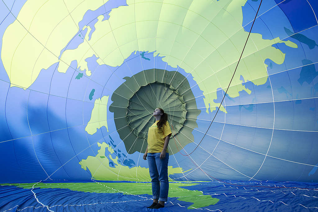 A hot-air balloon with a globe design, a woman in yellow tshirt and jeans stands in front looking up 