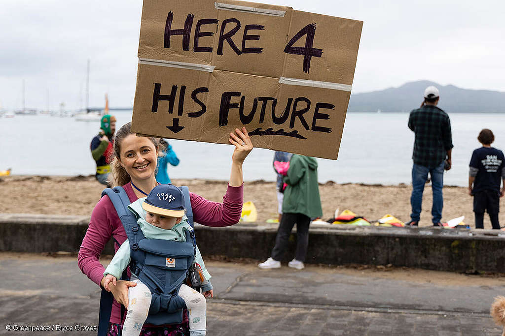 A woman with a baby in a front sling holds a sign saying 'Here for His Future' with the beach and Rangitoto in the background.