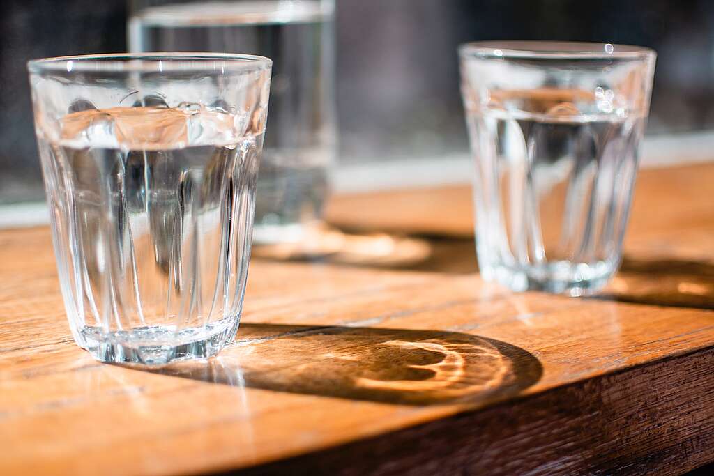 Three clear glasses of water on top of a wooden table, with sunlight behind.