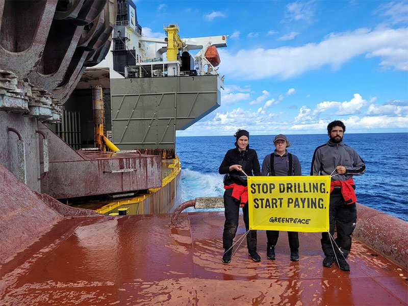 Three protesters from Greenpeace UK stand on the desck of a Shell oil platform and hold a large yellow banner with the words Stop drilling. Start paying. Three protesters from Greenpeace UK stand on the desck of a Shell oil platform and hold a large yellow banner with the words Stop drilling. Start paying.