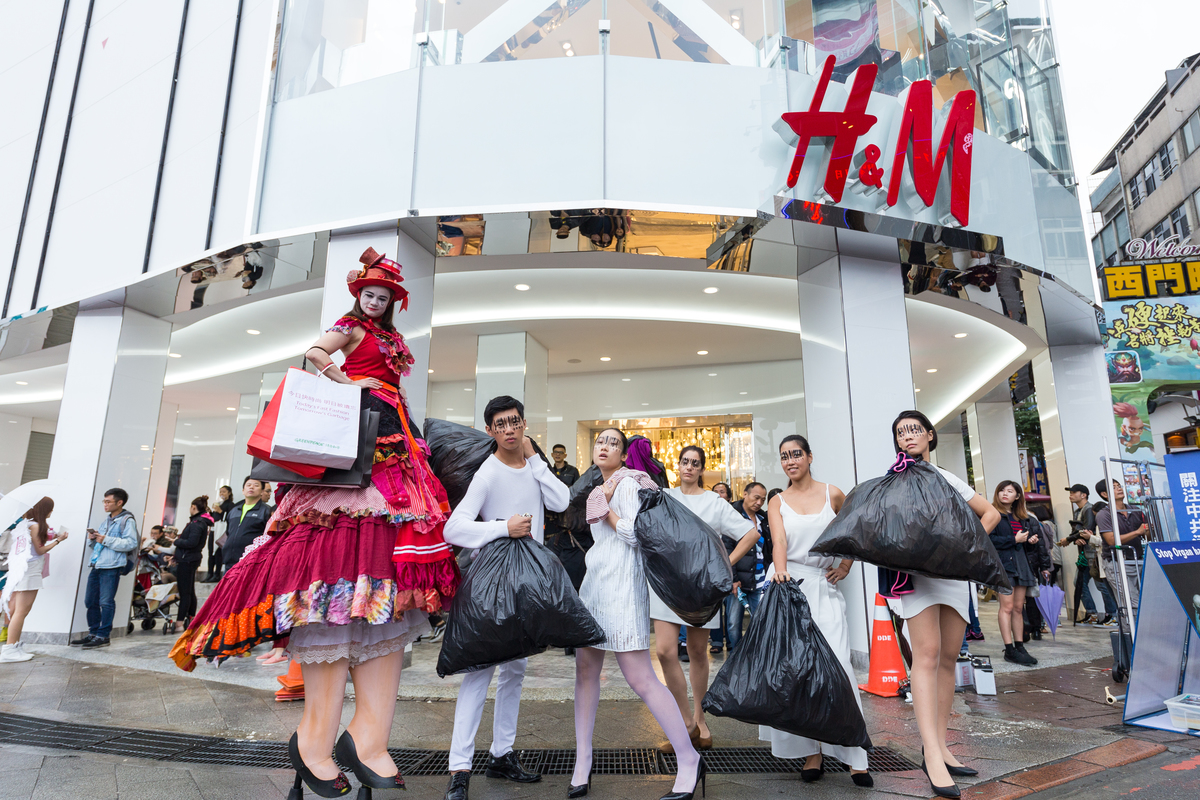 Street performance against fast fashion outside an H&M store with a person on stilts and people carrying black rubbish bags