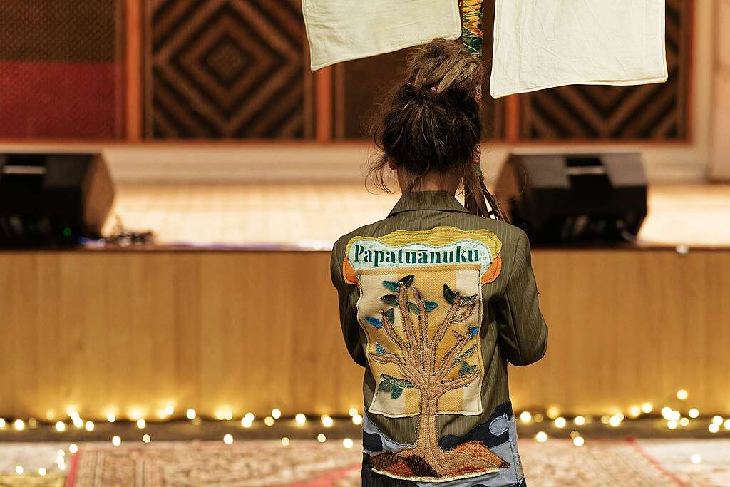 Someone facing away has a jacket with a beautiful stitched design on the back of a tree and the word Papatūānuku