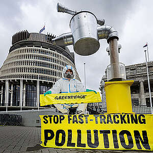 A greenpeace actvist wearing a hazmat suit and mask sits atop a giant effluent tap in front of parliament. The tap is filling a large drinking glass for the Prime Minister with cow effluent and a banner reads: Stop Fast-tracking Pollution