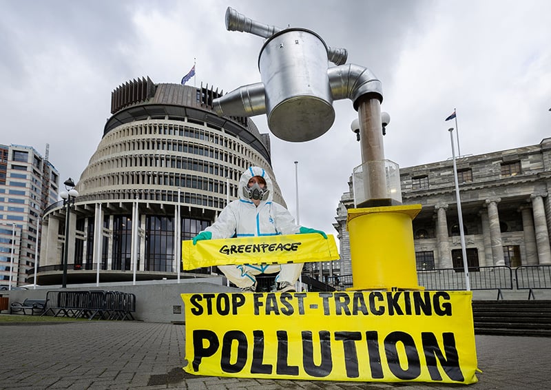 A greenpeace actvist wearing a hazmat suit and mask sits atop a giant effluent tap in front of parliament. The tap is filling a large drinking glass for the Prime Minister with cow effluent and a banner reads: Stop Fast-tracking Pollution