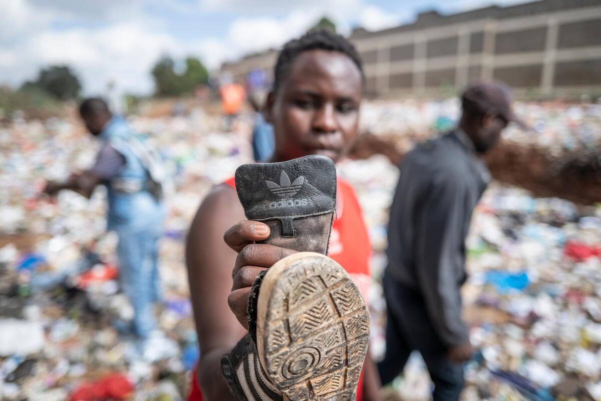 A person, out of focus, holds an old Adidas shoe, in focus. In the background are two people in a landfill. Fast Fashion Research in Kenya. © Kevin McElvaney / Greenpeace