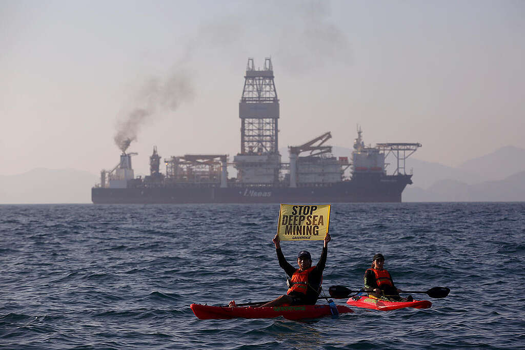 Two Greenpeace activists in kayaks hold a sign saying "Stop Deep Sea Mining". Behind them, is a deep sea mining vessel. Two Greenpeace activists in kayaks hold a sign saying "Stop Deep Sea Mining". Behind them, is a deep sea mining vessel.