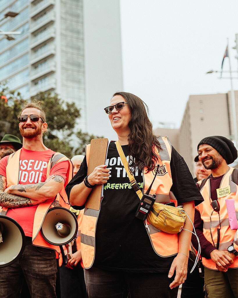 The Greenpeace volunteer coordinator, Charlie, at a protest on Queen Street. She is surrounded by other volunteers, all wearing orange safety vests.