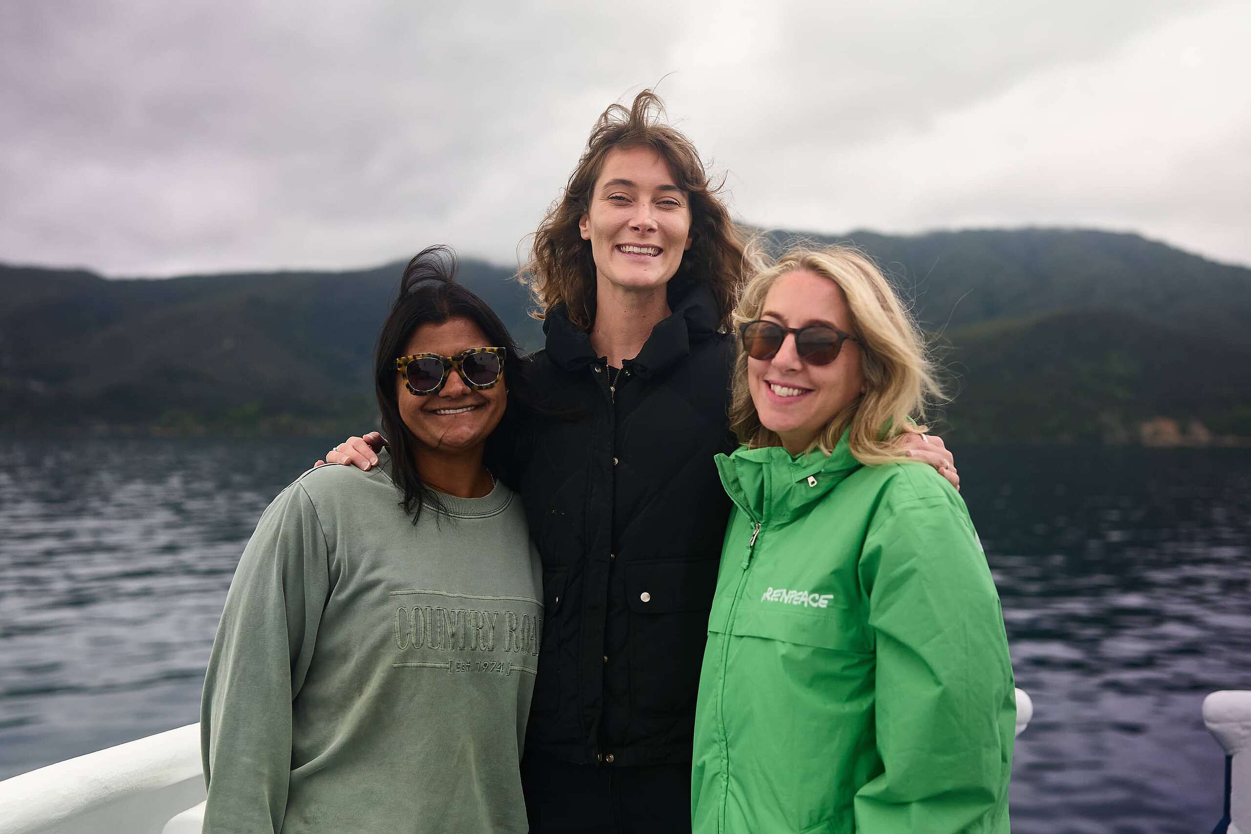 Team members Kat Goddard, Paige Maroni and Ellie Hooper onboard the Seamount Expedition stand against the rail outside with land in the background