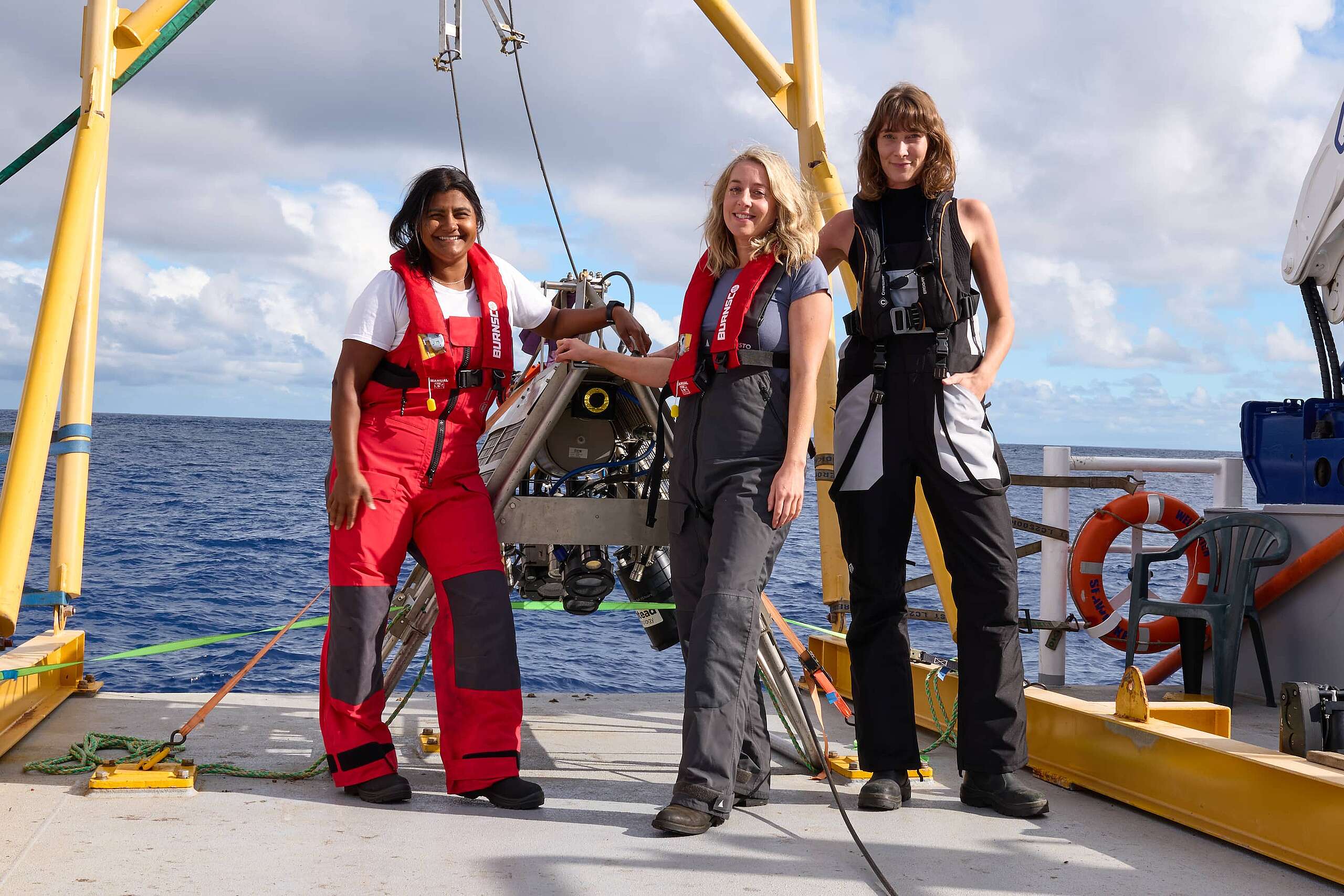 Three members of the Greenpeace Seamounts Expedition stand on the deck of the specialist research vessel looking at the camera with the ocean behind them. From left to right Kat Goddard, lead researcher, Ellie Hooper, oceans campaigner, Dr Paige Maroni, polar and deep-sea molecular biologist