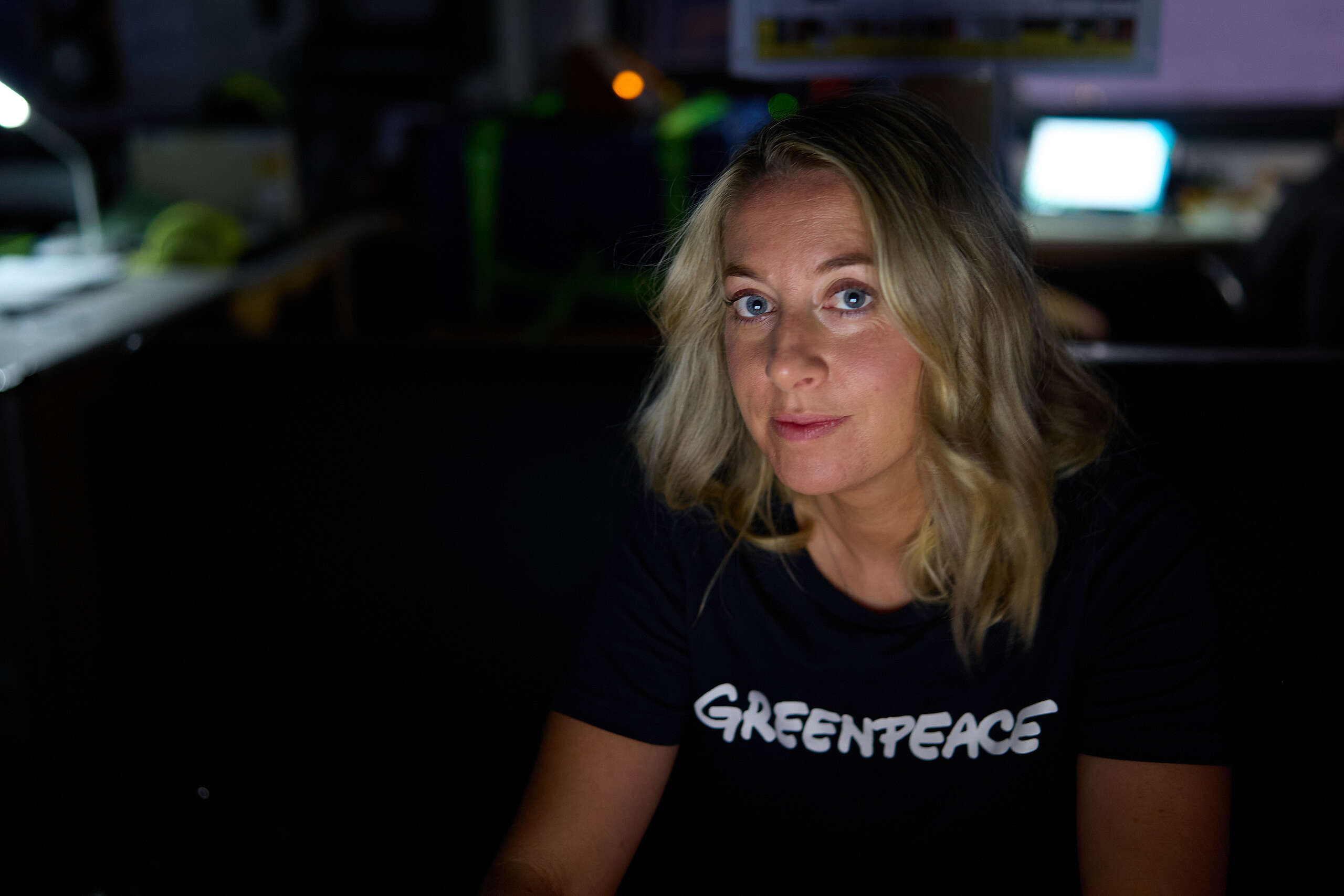 Greenpeace campaigner Ellie Hooper, wearing a t-shirt with Greenpeace on it, looks at the camera with light from a computer screen lighting her face