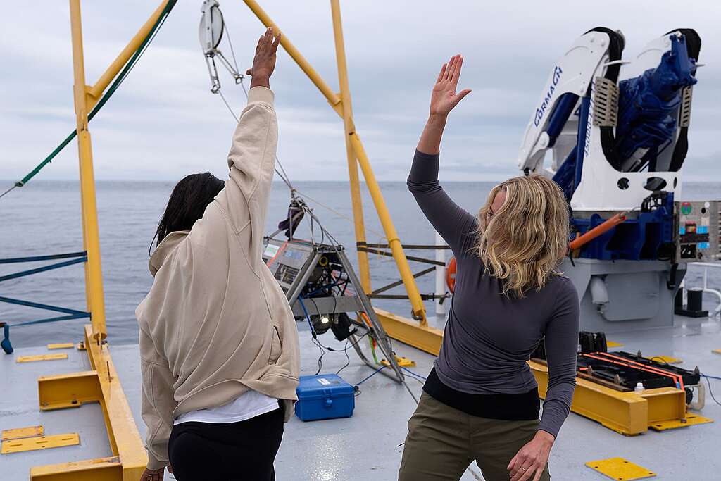 Lead scientist Kat Goddard stands on the left, right arm raised ready to high five Ellie Hooper, expedition lead, to her left. Both look happy. Lead scientist Kat Goddard stands on the left, right arm raised ready to high five Ellie Hooper, expedition lead, to her left. Both look happy.