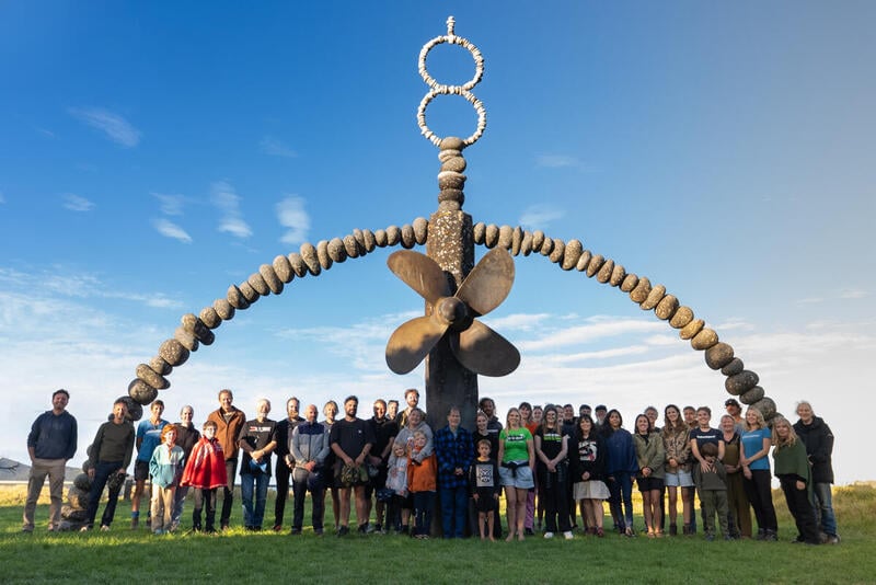 A line of people standing under the Rainbow Warrior memorial at Matauri Bay, with a rainbow made of stones and the ship propellor A line of people standing under the Rainbow Warrior memorial at Matauri Bay, with a rainbow made of stones and the ship propellor