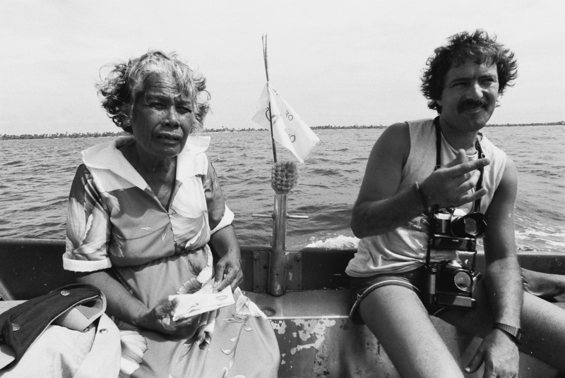 Two people sitting in the back of a small boat, the man on the right has a camera round his neck