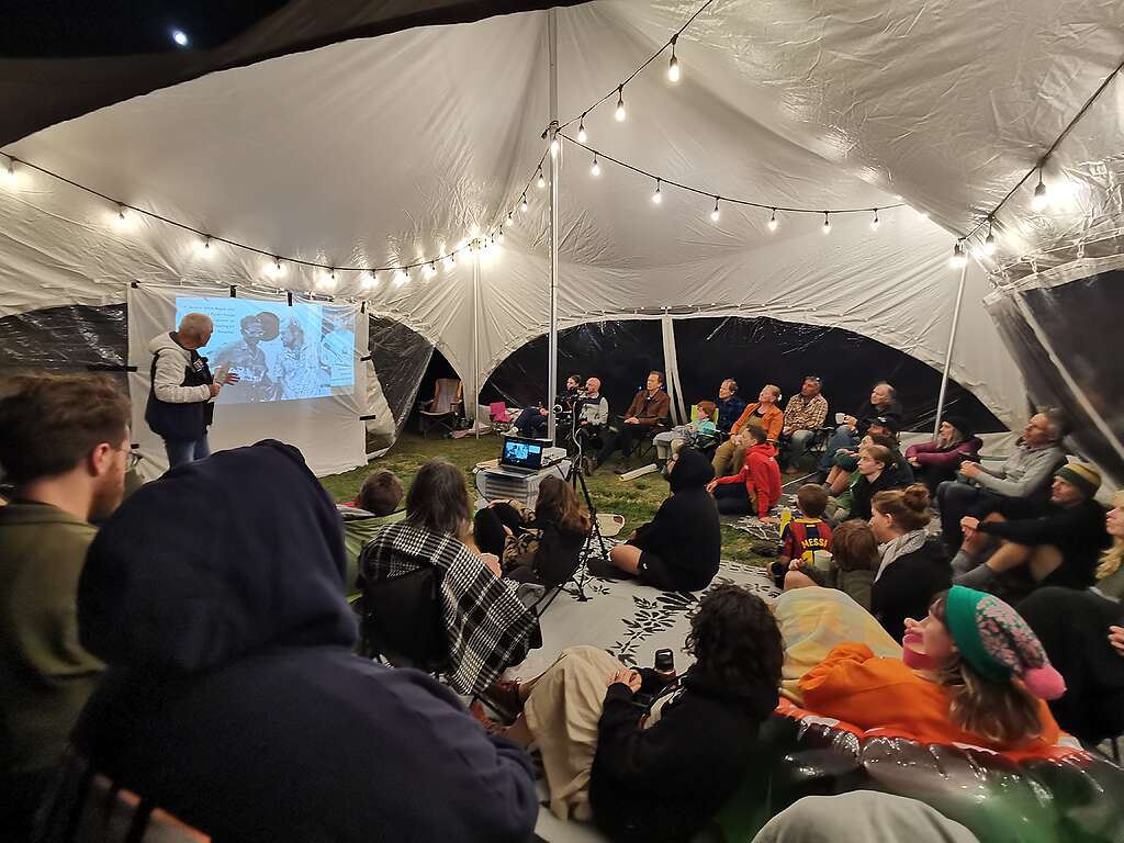 A group of people under a marquee with lights at night are watching a presentation.