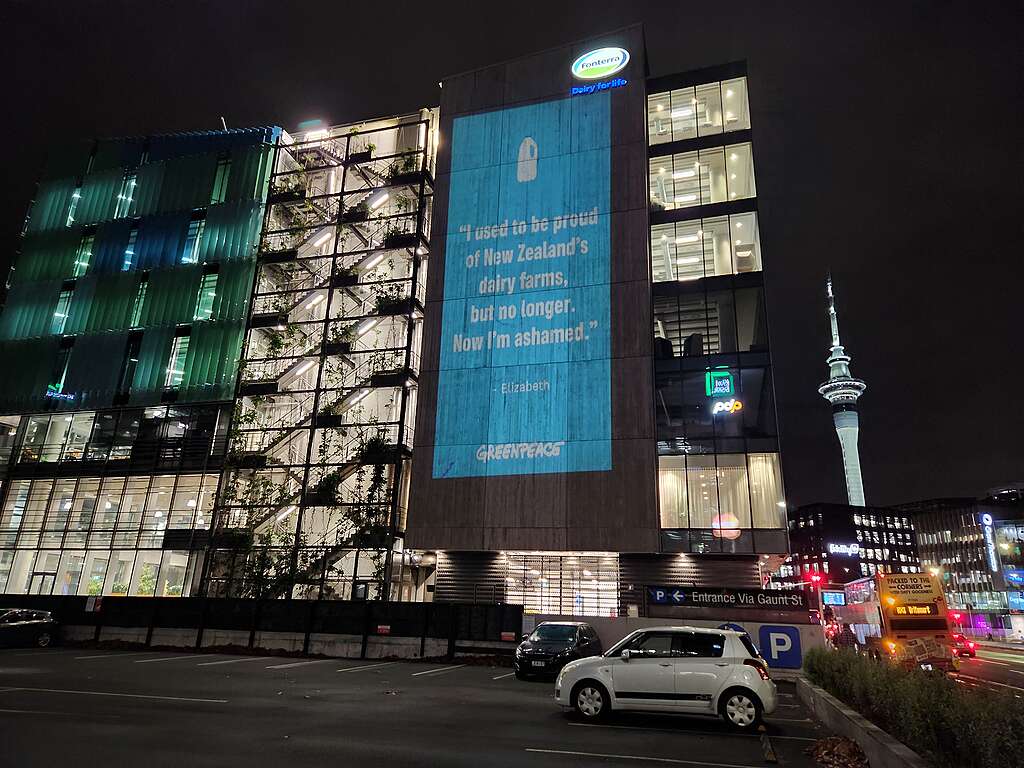 Greenpeace protests Fonterra environmental pollution by projecting onto their Auckland HQ. The projection is a blue graphic with white text. A quote reads 'I used to be proud of New Zealand's dairy farms, but no longer. Now I'm ashamed." The quote is attributed to Elizabeth, and the graphic takes up the whole wall of the multi-story building.