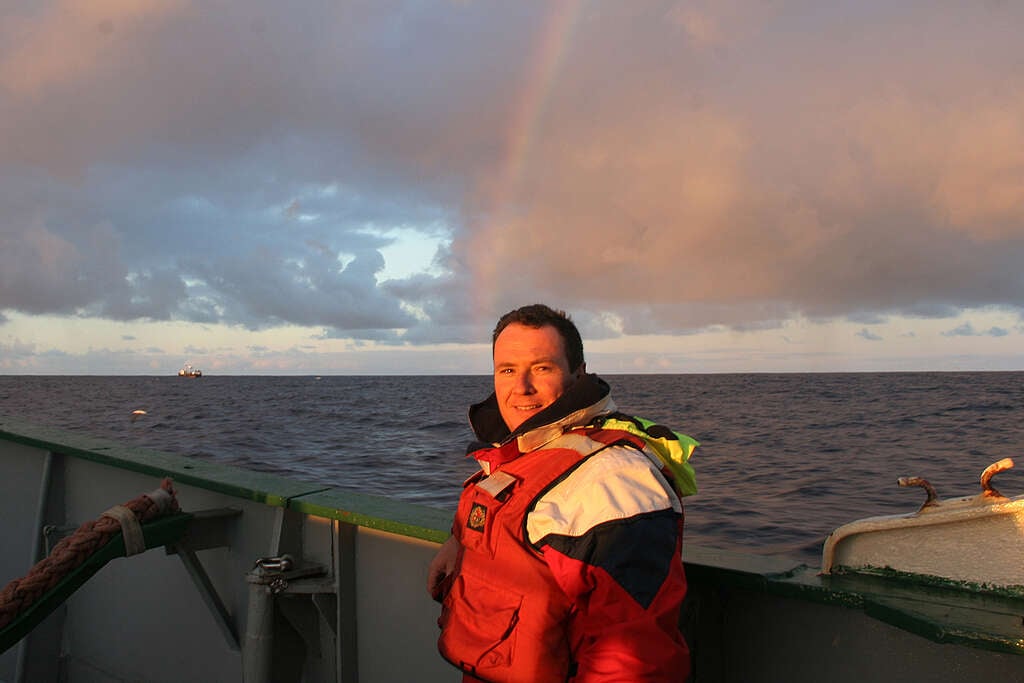 An image of the videographer on the 2005 Rainbow Warrior voyage, Wooly, who captured the moment the coral was brought up from the deep on video. Wooly stands on the deck of the Rainbow Warrior by the railing, wearing a red vest. There is a rainbow in the background, and far in the distance is a trawl vessel.