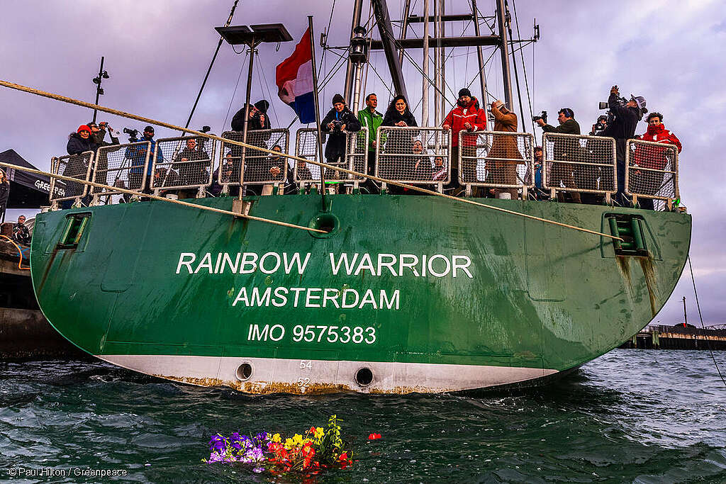 People stand at the back of the Rainbow Warrior ship, as flowers float on the surface below People stand at the back of the Rainbow Warrior ship, as flowers float on the surface below