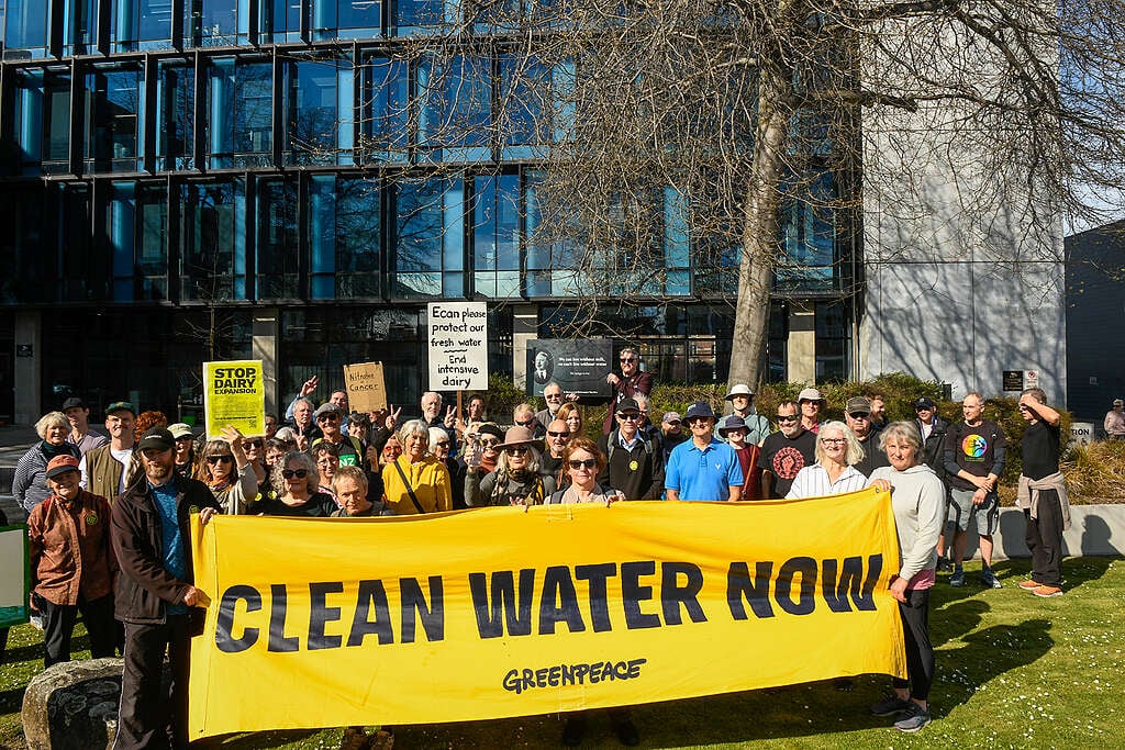 Frustrated Cantabrians rally outside Environment Canterbury's Christchurch HQ before a declaration of nitrate emergency. A crowd of 50-60 people stand behind a yellow banner which reads 'Clean Water Now' in black text. Other members of the crowd are holding signs calling on ECan to protect fresh water and stop dairy expansion.