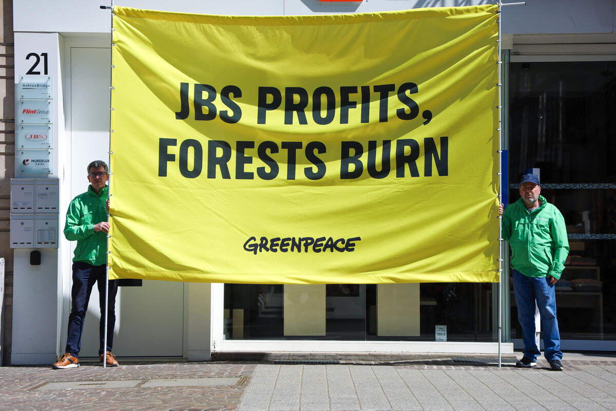 Greenpeace activists protest against JBS in Luxembourg. Two activists in green raincoats hold a large banner which reads 'JBS profits, forests burn'.
