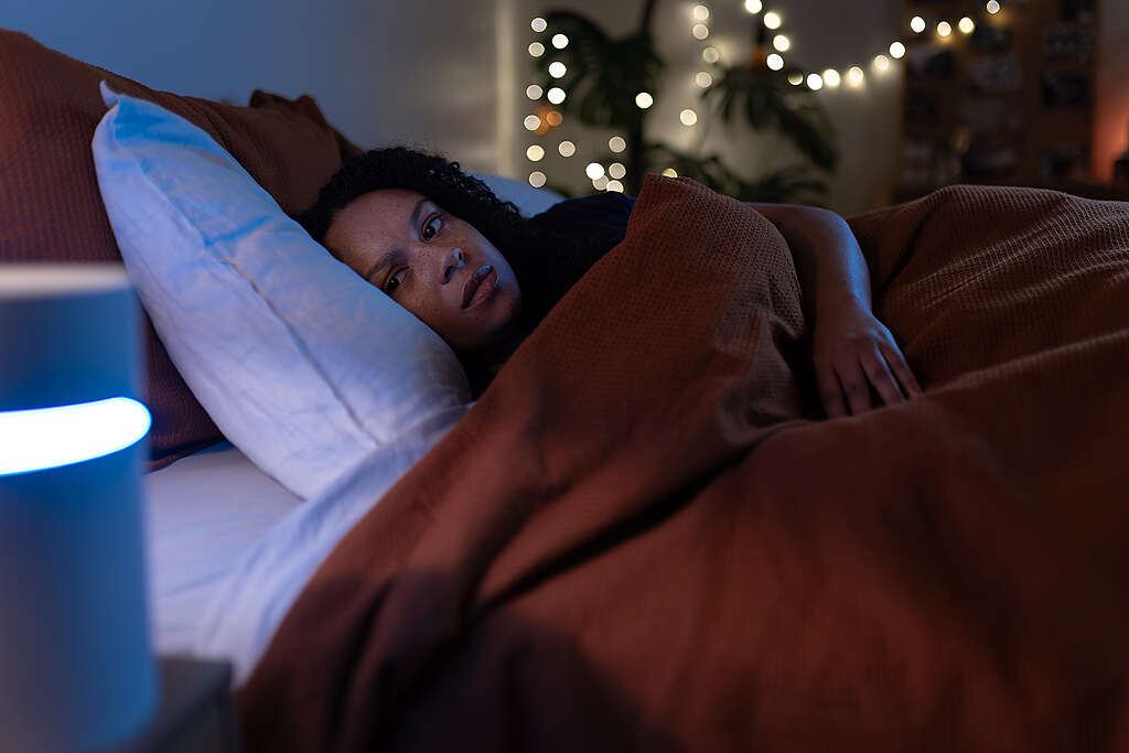 Young woman lying in bed looking at an electronic virtual assistant device with a look of concern on her face.