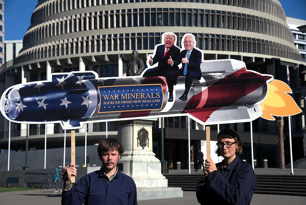 Two people in blue hold above their heads a sign in the shape of a missile with Trump and Luxon riding