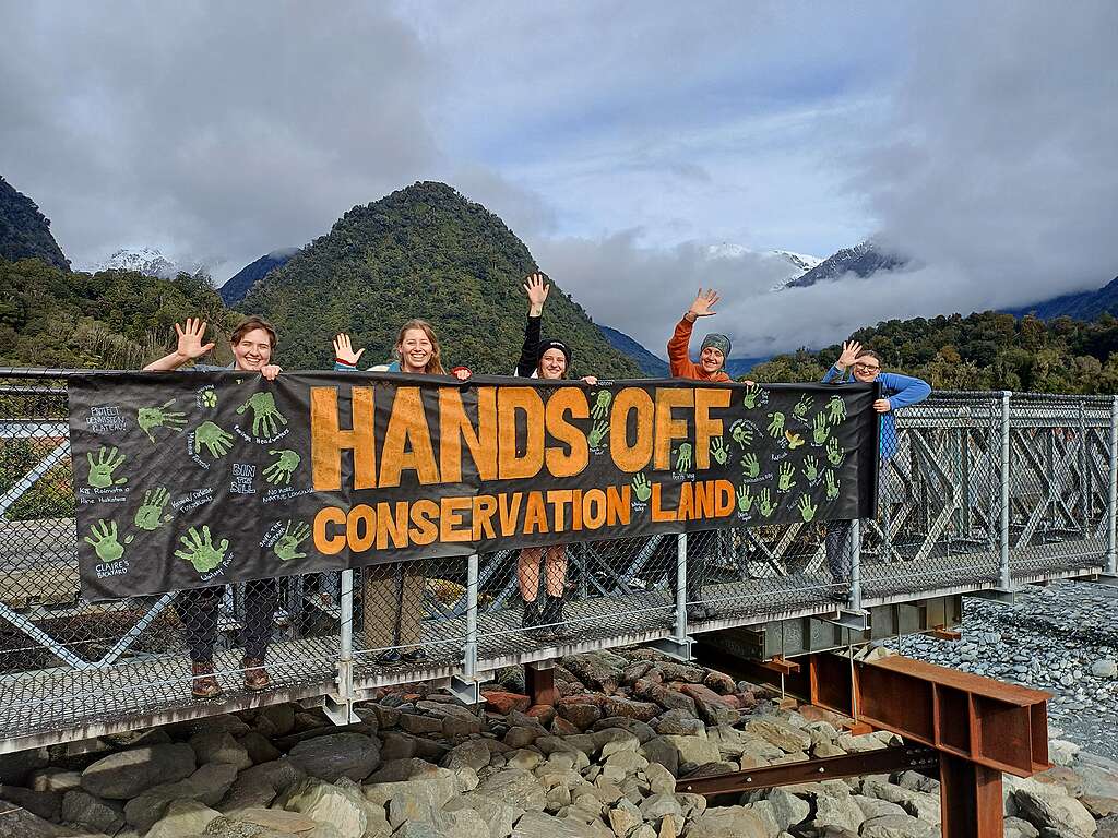 People on a walking bridge with mountain background hold a a hand made banner: HANDS OFF CONSERVATION LAND