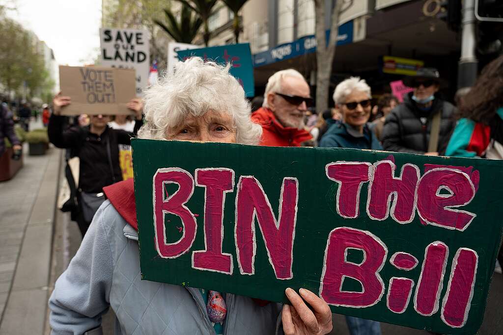 A woman with white hair looks from behind a sign: BIN THE BILL
