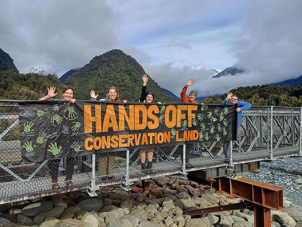 5 people stand on a walking bridge and wave, in front of beautiful mountain scenery. A hand made banner says HANDS OFF CONSERVATION LAND 