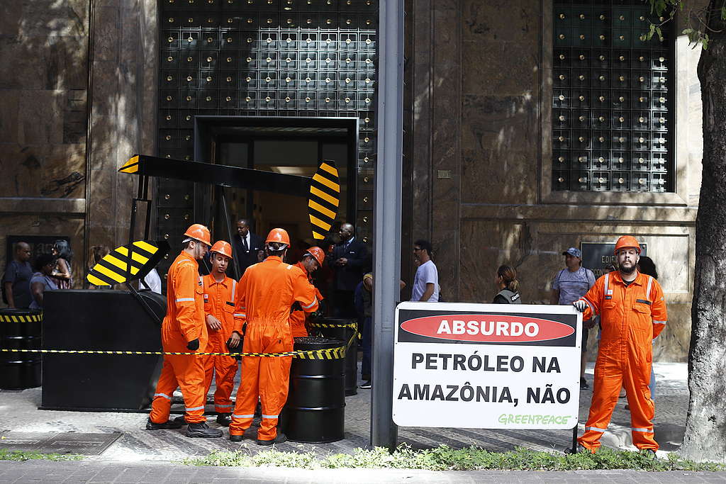 pessoas simulam a exploração de petróleo com uma máquina e barris em frente ao prédio do governo. 