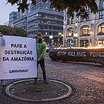 Ativistas do Greenpeace Alemanha mostram cartaz com a frase "pare a destruição da Amazônia"; tronco de árvore com a frase em inglês "stop killing forests" queima ao fundo