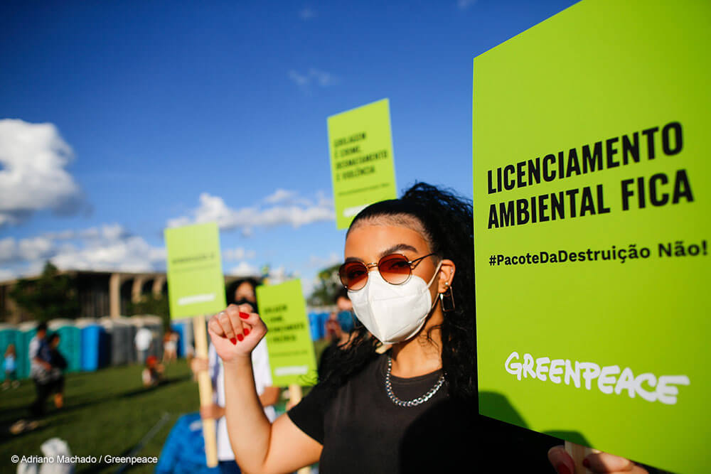 Imagem de uma jovem de cabelos castanhos e óculos escuros participa do Ato Pela Terra, em Brasília. Ela tem o punho direito erguido e, na outra mão, segura uma placa verde com a mensagem "Licenciamento ambiental fica"