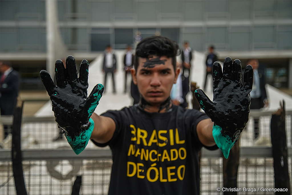Foto de um homem em uma ação direta não violenta executada na frente do congresso nacional. Suas mãos estão sujas de um material similar ao petróleo e sua camisa diz "Brasil Manchado de Óleo", em alusão à uma contaminação de petróleo ocorrida em uma praia brasileira em 2021.