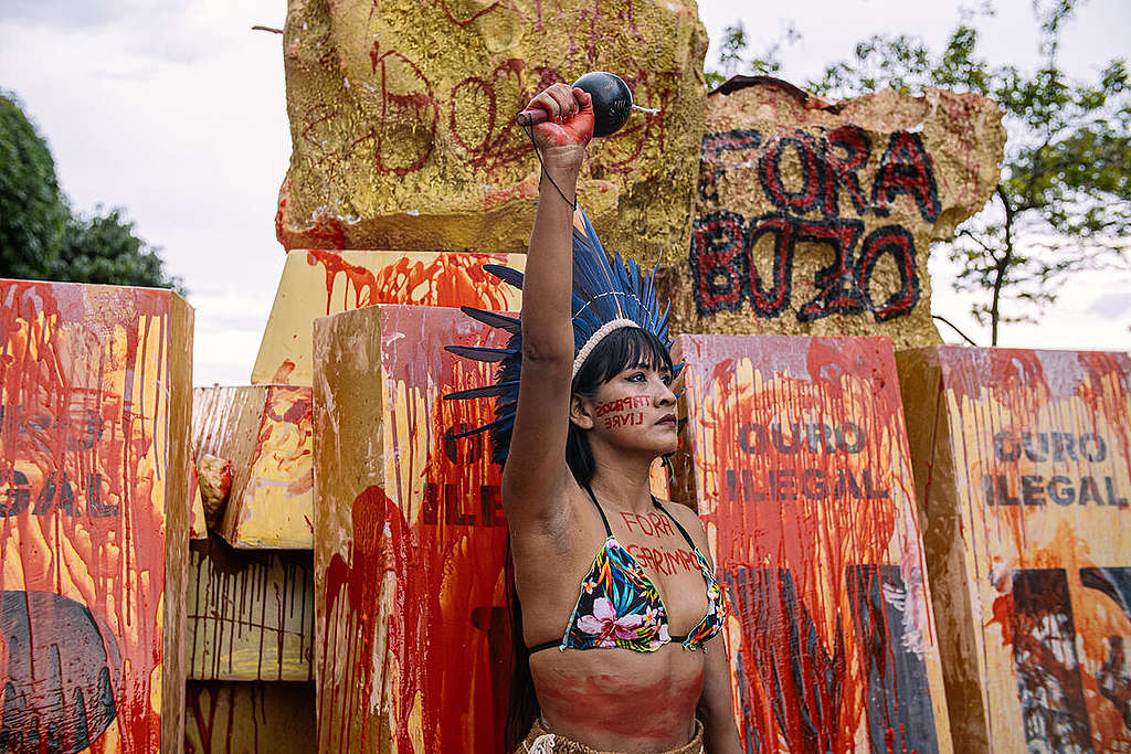 Mulher indígena com o punho erguido em protesto contra o garimpo ilegal. Ela usa um cocar azul e tem a frase "fora garimpo" escrita em seu corpo com uma tinta vermelha, simulando sangue.