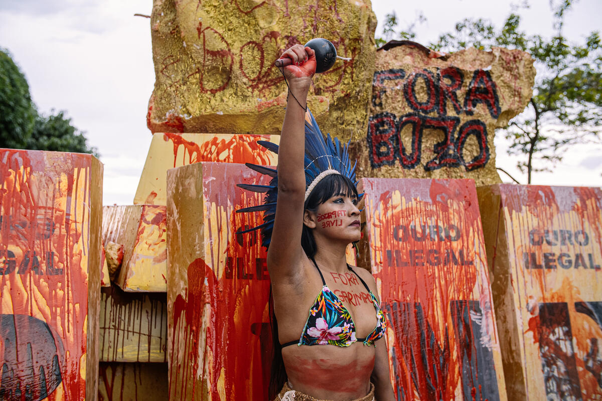 Mulher indígena com o punho erguido em protesto contra o garimpo ilegal. Ela usa um cocar azul e tem a frase "fora garimpo" escrita em seu corpo com uma tinta vermelha, simulando sangue.