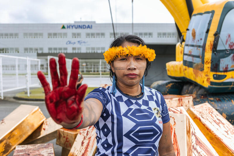 mulher indígena com a mão suja de sangue cenográfico, na frente da fábrica da Hyundai