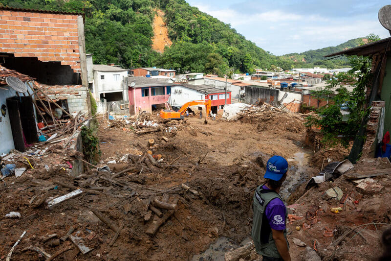 Vista do alto, em primeiro plano, lama sobre casas soterradas no litoral norte de São Paulo e voluntário do Greenpeace Bertioga identificado com colete e boné com o logo da organização está de costas. No horizonte, algumas casas destruídas, mas ainda de pé. E ao fundo, morros com vegetação, mas com aberturas marcadas por buracos de terra que derrubaram as árvores após deslizamentos.