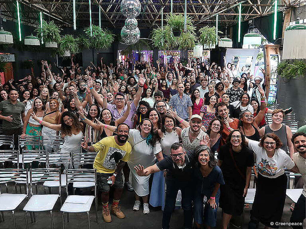 Fotografia retrata coletivo de pessoas em um espaço com cadeiras. Elas estão com os braços levantados e sorrindo. Imagem feita durante  confraternização de fim de ano do Greenpeace Brasil em 2022.