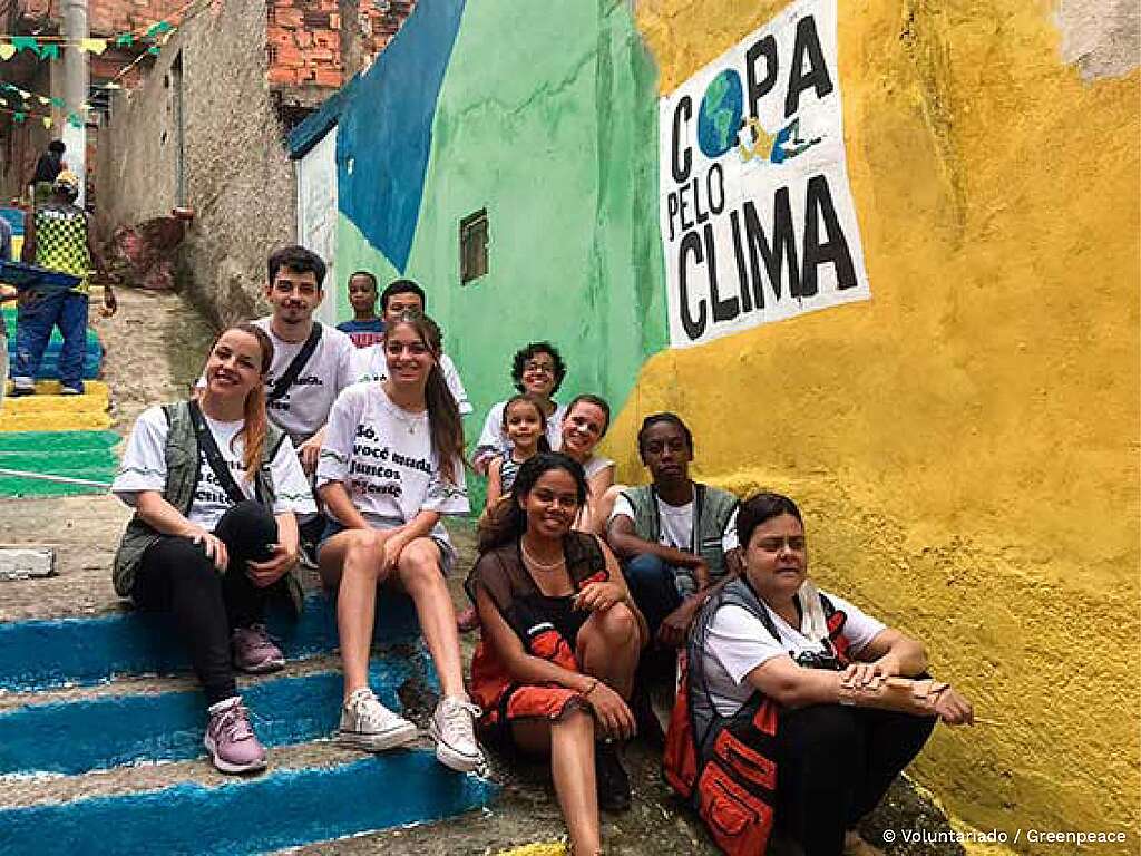 Fotografia de jovens voluntários do Greenpeace Brasil juntos, sentados em uma escada. Ao lado direito, um cartaz com a frase: Copa Pelo Clima grudado em uma parede amarela