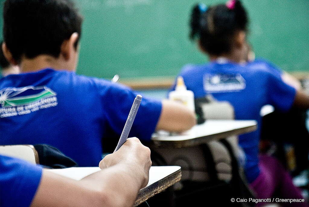Em um plano aberto ambientado em uma sala de aula de uma escola de educação infantil, mostra-se crianças sentadas em carteiras, escrevendo.