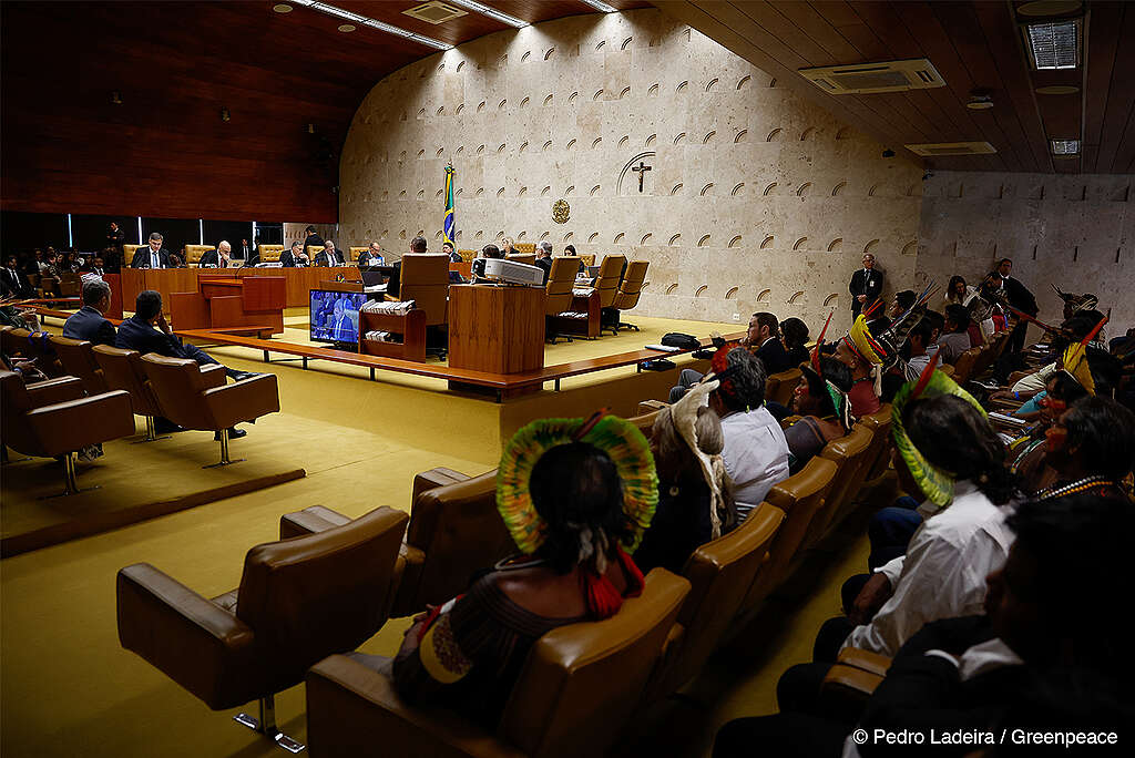 Em um plano aberto, a imagem mostra o momento do julgamento da tese do Marco Temporal. Ao lado esquerdo temos os juízes exercendo seus votos e ao lado direito uma plateia, de maioria indígena, acompanhando a votação.