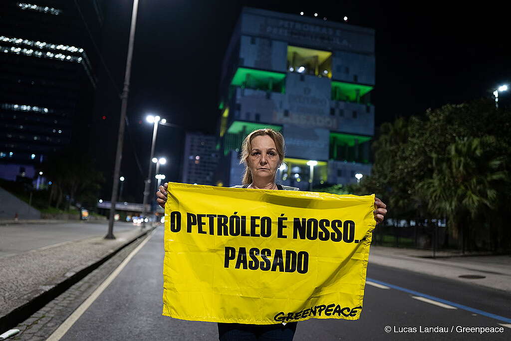 Em primeiro plano há uma ativista do Greenpeace segurando um cartaz com os dizeres "O petróleo é nosso passado". Ao fundo temos o prédio da Petróbras, no Rio de Janeiro, e nele os ativistas projetam mensagens em uma ação que pede a transição energética justa e também a não exploração de petróleo na Amazônia.