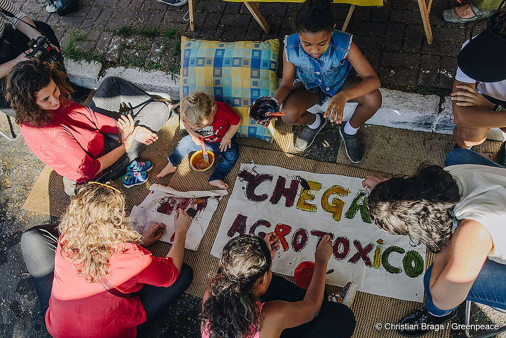 Em plano médio, numa imagem aérea, voluntários do Greenpeace realizam atividades de ativismo e alimentação para crianças e adultos, como jogos e oficinas de produção de tinta natural e cartazes com os dizeres "Chega de Agrotóxicos".