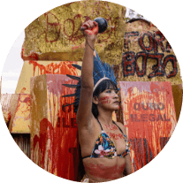 Foto de uma mulher indígena com a mão levantada em uma ação direta não violenta contra o garimpo. Ao fundo temos representações de barras de ouro marcadas pelo sangue dos povos originários.