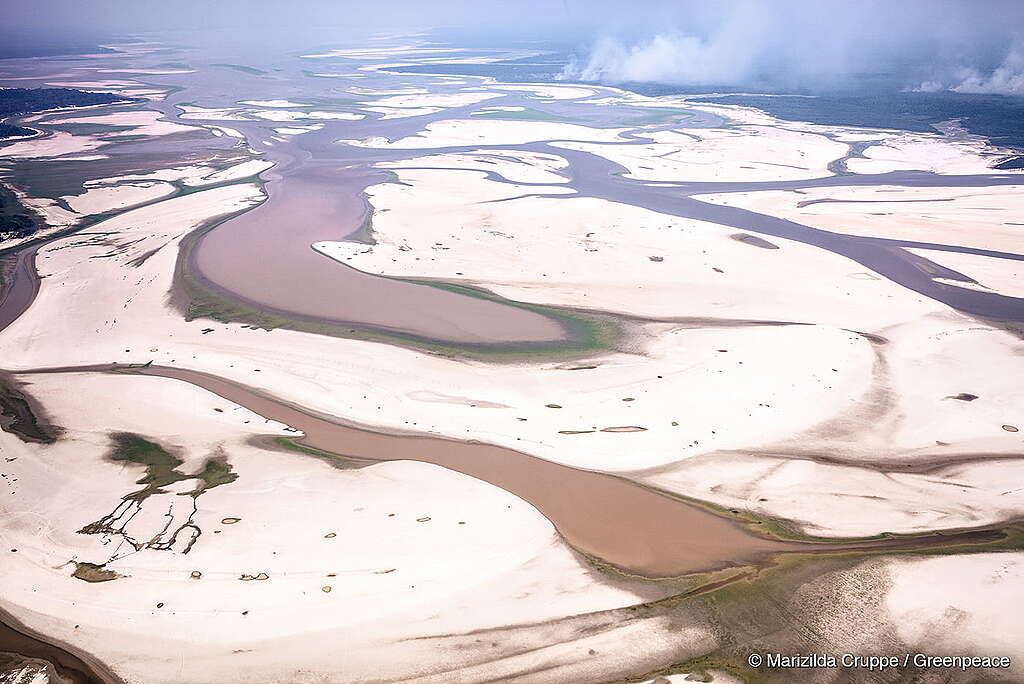 A fotografia aérea mostra o impacto devastador da seca na Amazônia, com rios e lagos secando, isolando comunidades e causando uma grave emergência ambiental. Ao fundo, temos uma queimada de grandes proporções.