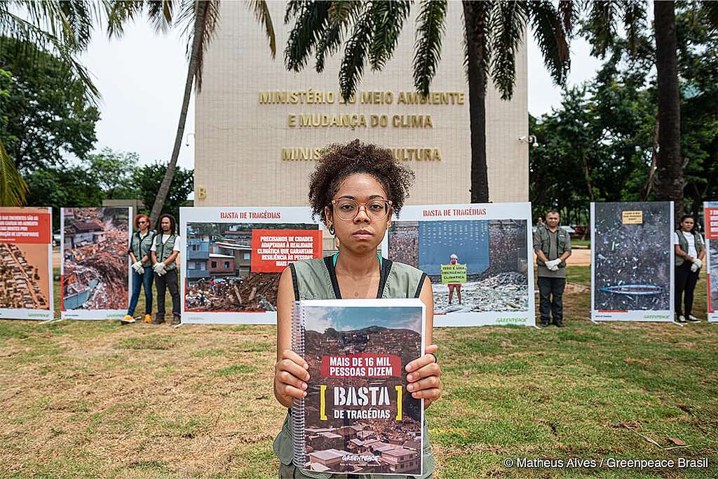 A foto mostra uma mulher em frente ao ministério do meio ambiente e mudança do clima segurando um caderno com as assinaturas do abaixo-assinado Basta de Tragédias. 

A mulher é negra, possui cabelos cacheados e usa óculos. Ela veste o colete do Greenpeace. Ao fundo temos uma exposição de fotos do Greenpeace da campanha de justiça climática.
